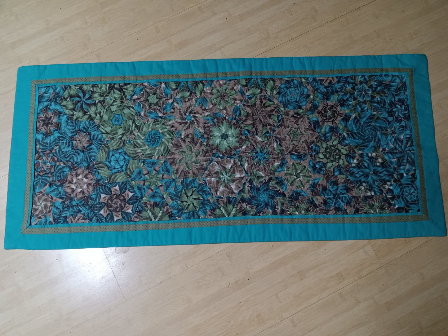A table runner made of blue, green, and brown fabric arranged in a kaleidoscope pattern. The table runner has a blue border and is on a wood panel background.