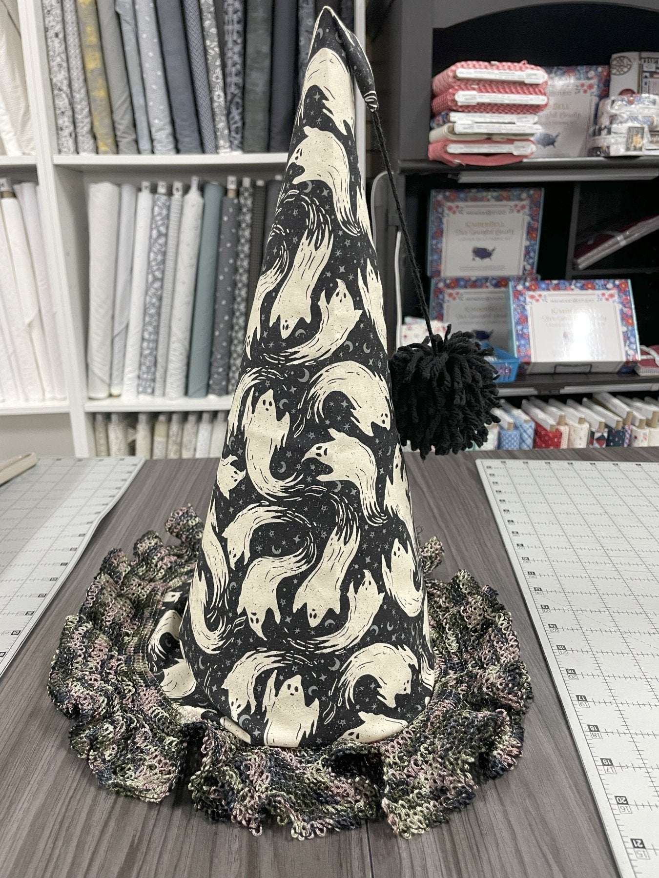 A tall witches hat made from black fabric with ghosts printed on it, and a black and grey lace trim for the brim. A black pompom is attached to the tip by string. The hat is sitting on a cutting table with bolts of fabric in the background.