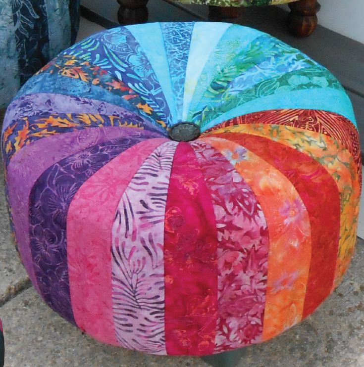 A circular tuffet or ottoman made of strips of Batik fabric arranged in a rainbow gradient from orange, to pink, purple, dark blue, light blue, and back to orange. It is sitting on a concrete floor.
