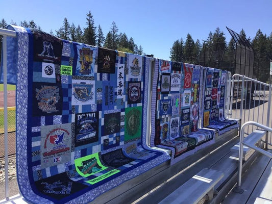 T-shirt quilt made from various shirts displayed on a fence with a clear blue sky and trees in the background.