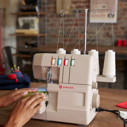 Hands guiding green fabric under the presser foot of a Singer HD0420 serger with four thread cones