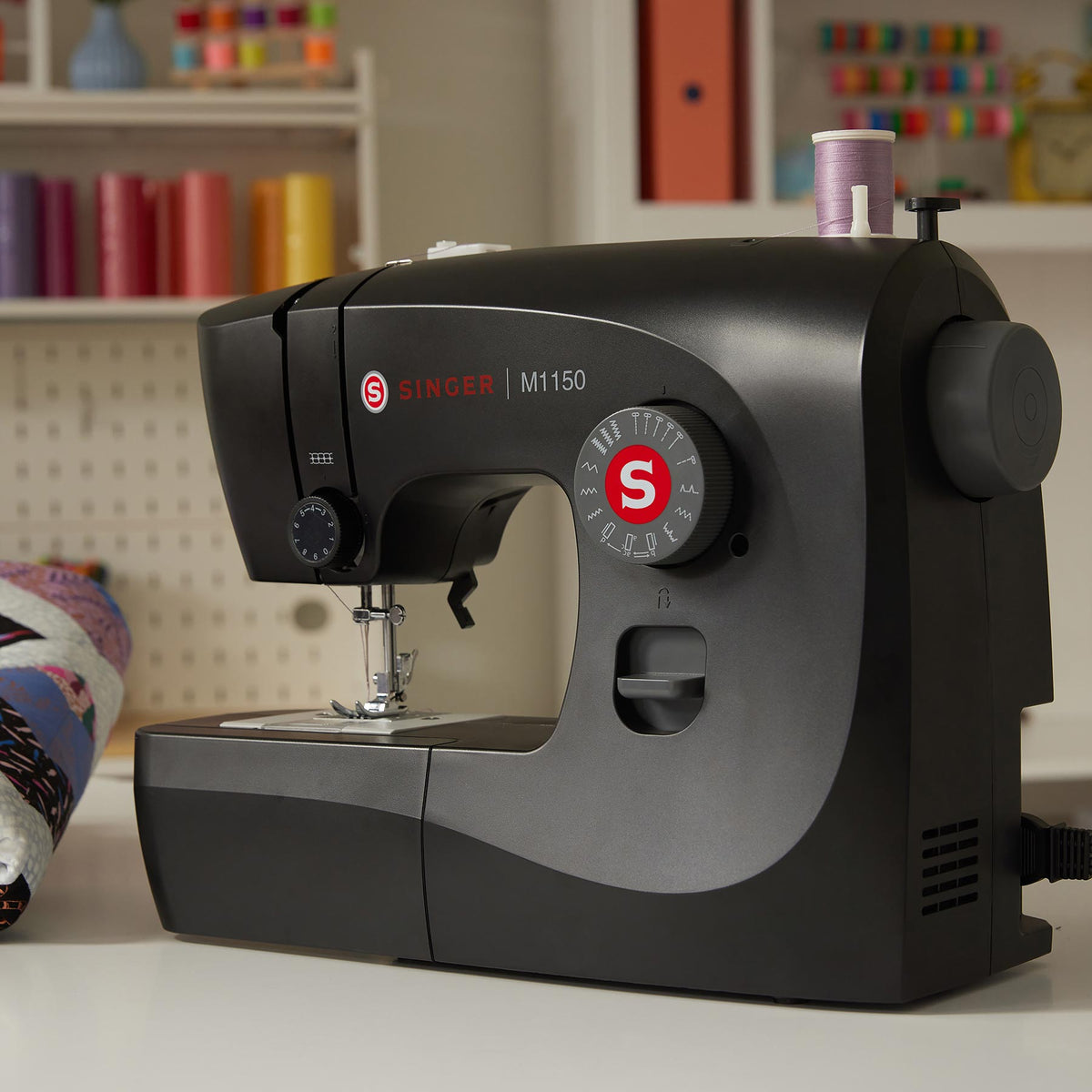 Singer M1150 Black sewing machine on a table beside spools of thread and a quilt