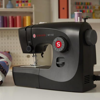 Singer M1150 Black sewing machine on a table beside spools of thread and a quilt