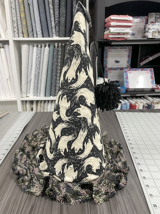 A tall witches hat made from black fabric with ghosts printed on it, and a black and grey lace trim for the brim. A black pompom is attached to the tip by string. The hat is sitting on a cutting table with bolts of fabric in the background.
