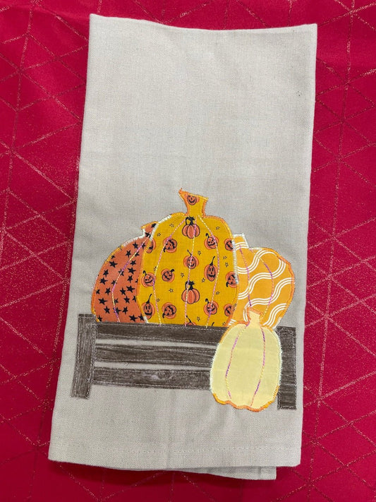 A grey tea towel with an applique design of three pumpkins sitting on a wooden fence, and a fourth pumpkin sitting in front of the fence to the right. The towel is on red background.