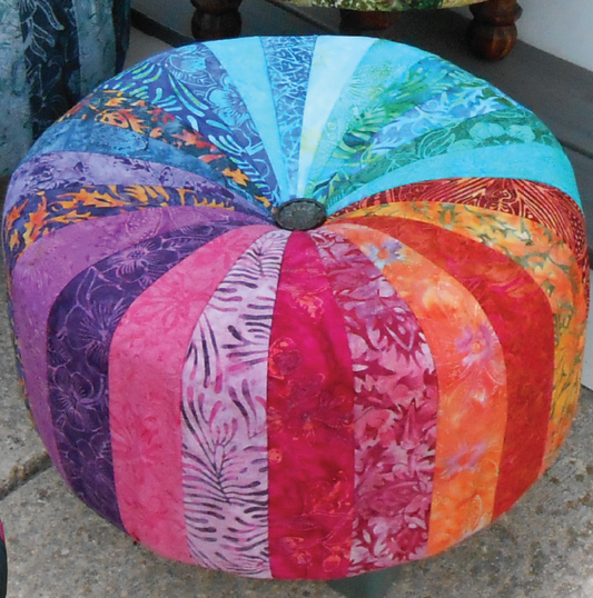 A circular tuffet or ottoman made of strips of Batik fabric arranged in a rainbow gradient from orange, to pink, purple, dark blue, light blue, and back to orange. It is sitting on a concrete floor.
