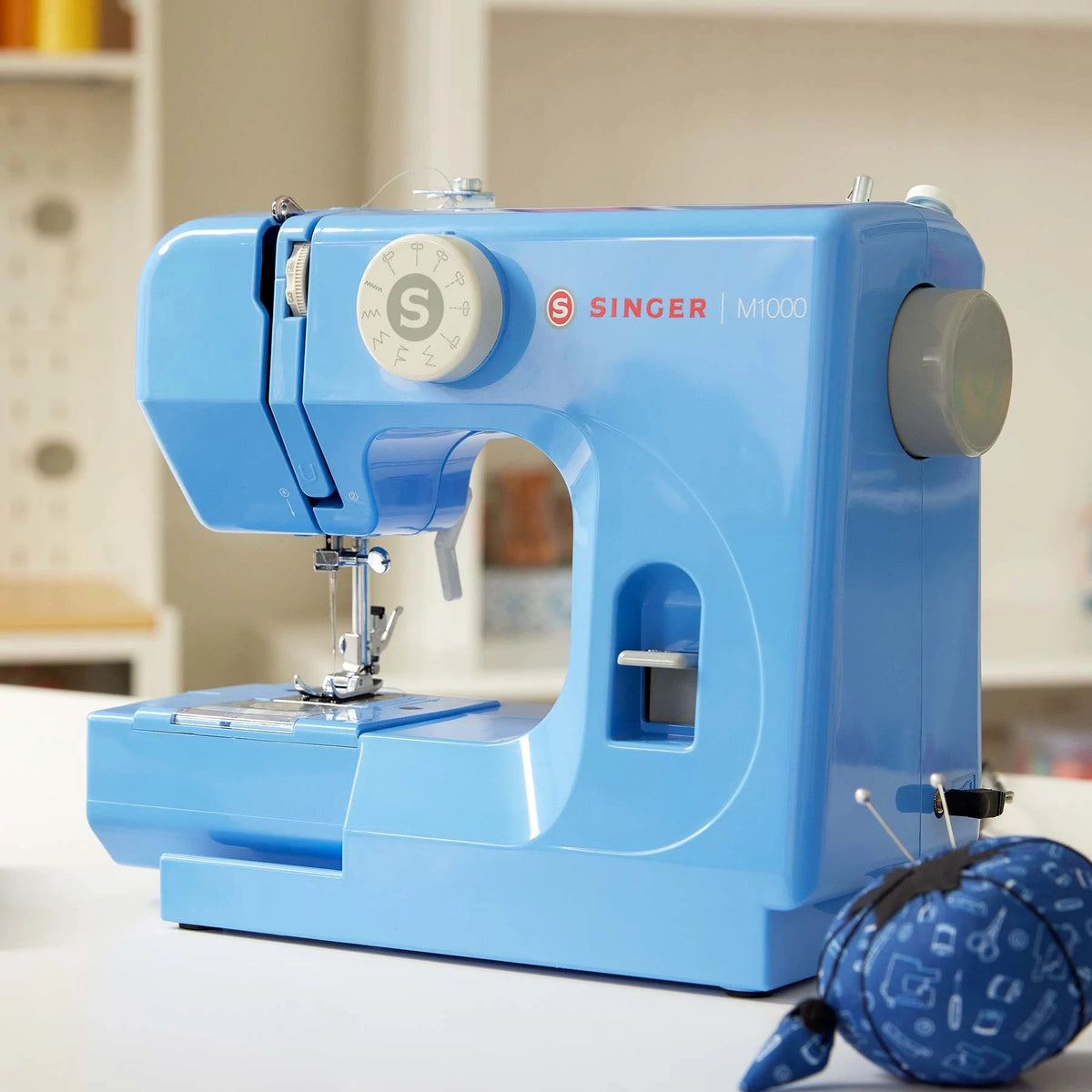 Singer M1000 Blue mending machine on a sewing table beside a pin cushion